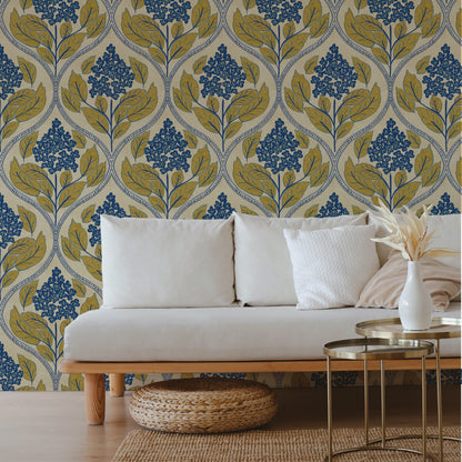 A styled living room scene featuring a white sofa with white pillows, a woven basket ottoman, and a white vase on gold side tables, set against a wall with a blue and olive green floral damask wallpaper pattern.