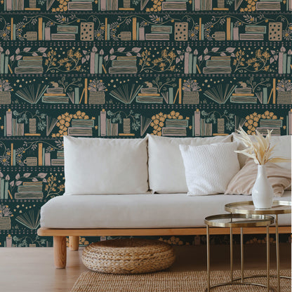 A styled living room scene featuring a white sofa with white and beige pillows, set against a dark teal wallpaper with a repeating pattern of books, flowers, and candles. In front of the sofa are two round gold-framed side tables with a white vase holding dried grass stems, and a woven round ottoman on the floor.