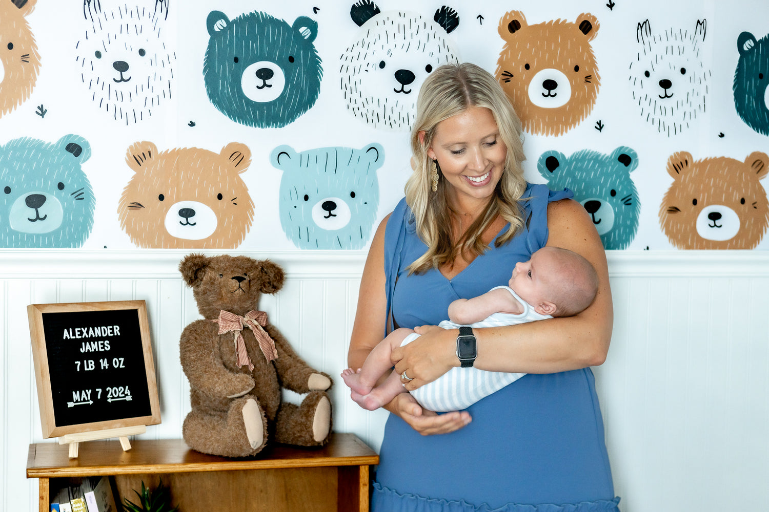 Woman holding a baby in front of a wall with bear illustrations and a teddy bear.