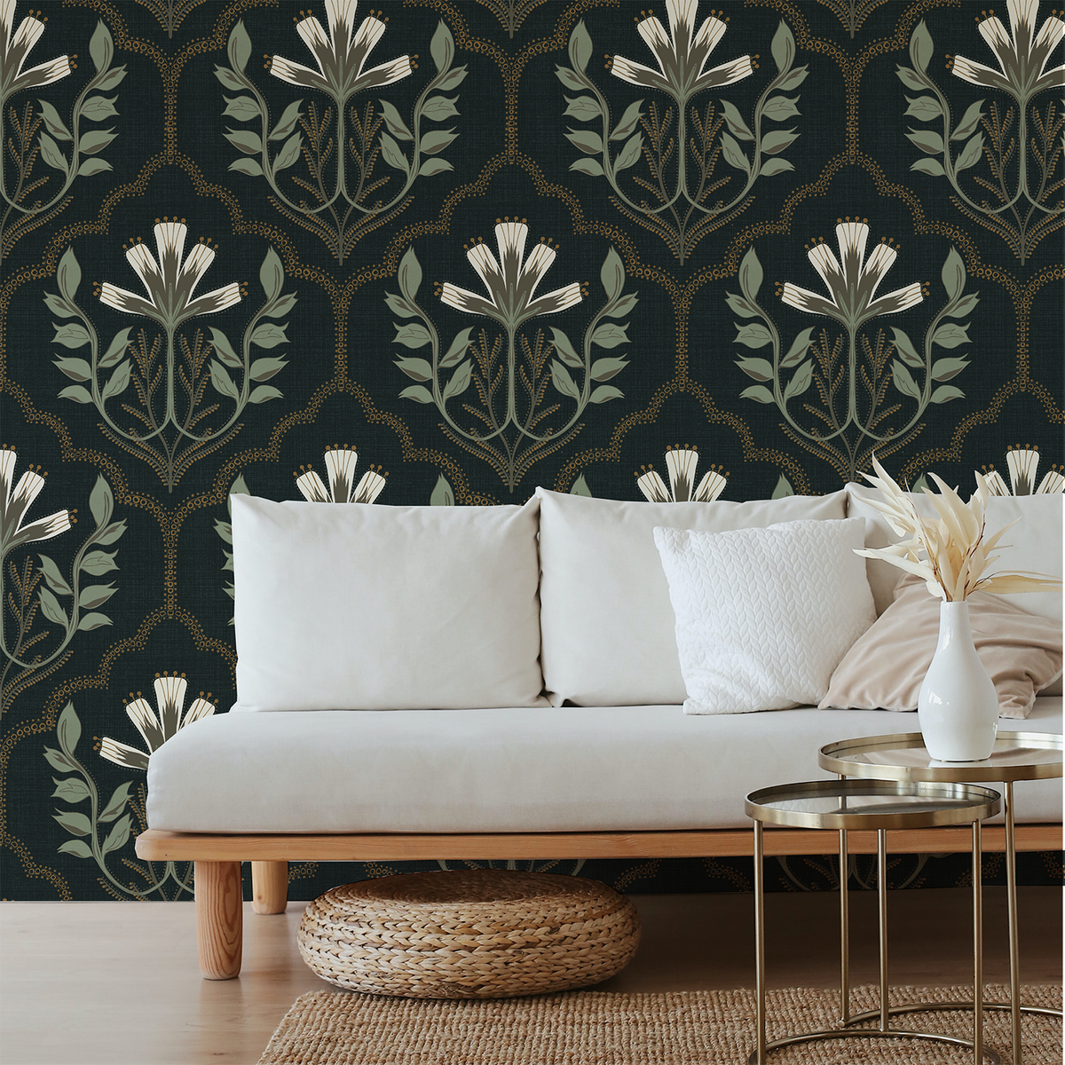 A styled living room scene featuring a white sofa against a dark green wallpaper with a repeating botanical damask pattern in muted green, beige, and gold. A white vase with dried grass sits on a gold metal side table in the foreground.