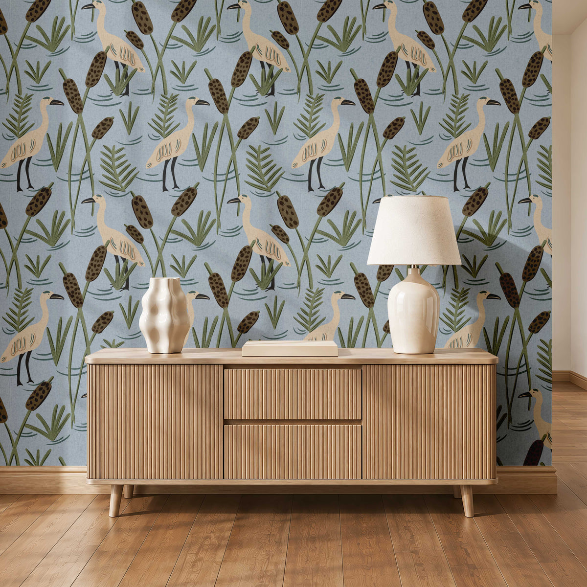 A styled interior scene showing a light wood sideboard against a blue wallpaper with a bird-and-reeds pattern, plus a white table lamp and decorative vases on top.