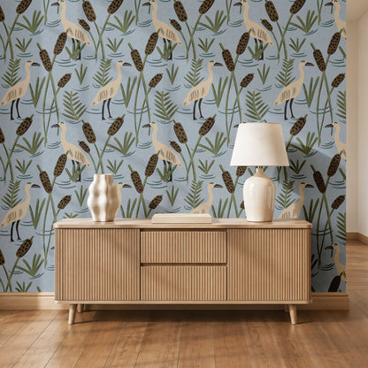 A styled interior scene showing a light wood sideboard against a blue wallpaper with a bird-and-reeds pattern, plus a white table lamp and decorative vases on top.