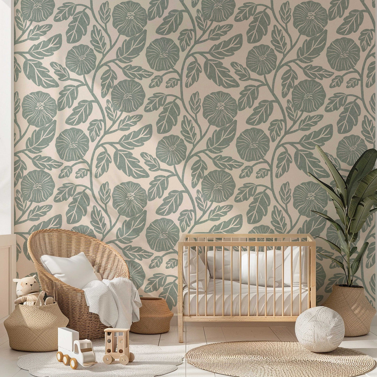 A styled nursery room featuring a wall covered in botanical wallpaper with a sage green leaf pattern on a warm beige background. The room is styled with a light wood crib, a woven wicker armchair with a white throw pillow, a potted plant, and various wooden toys on a light-colored floor.