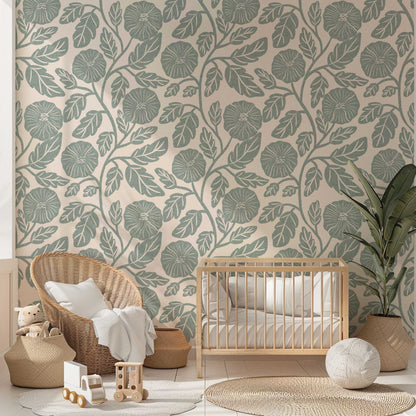 A styled nursery room featuring a wall covered in botanical wallpaper with a sage green leaf pattern on a warm beige background. The room is styled with a light wood crib, a woven wicker armchair with a white throw pillow, a potted plant, and various wooden toys on a light-colored floor.