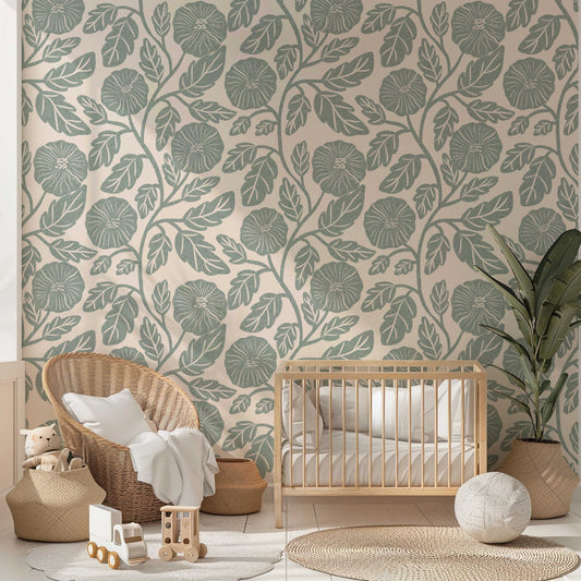 A styled nursery room featuring a wall covered in botanical wallpaper with a sage green leaf pattern on a warm beige background. The room is styled with a light wood crib, a woven wicker armchair with a white throw pillow, a potted plant, and various wooden toys on a light-colored floor.