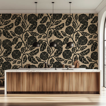 A modern kitchen interior featuring a bold black floral and leaf pattern wallpaper on a warm beige background, paired with a light wood cabinet island and black pendant lights.