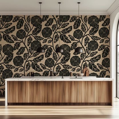 A modern kitchen interior featuring a bold black floral and leaf pattern wallpaper on a warm beige background, paired with a light wood cabinet island and black pendant lights.