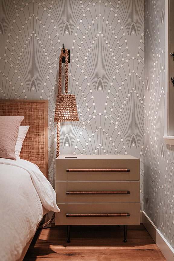 A styled bedroom corner featuring a beige three-drawer nightstand with copper-toned handles, set against a wall with white geometric wallpaper. A woven rattan headboard, white bedding, and a hanging woven pendant light are visible.