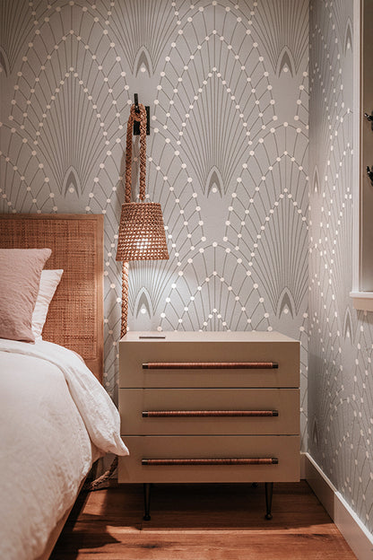 A styled bedroom corner featuring a beige three-drawer nightstand with copper-toned handles, set against a wall with white geometric wallpaper. A woven rattan headboard, white bedding, and a hanging woven pendant light are visible.