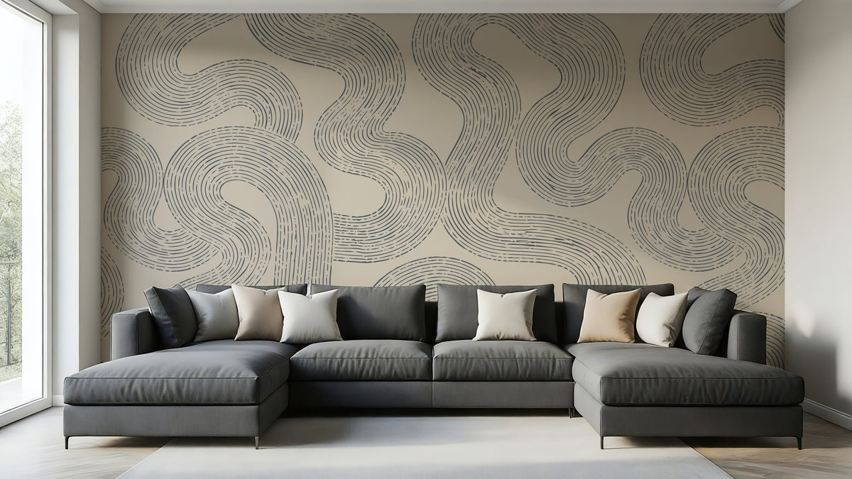 A modern gray sectional sofa with multiple throw pillows is positioned in front of a large wall covered in a beige wallpaper mural with a dark gray wavy line pattern. The room has light wood floors, a white window frame on the left, and a light gray rug on the floor.