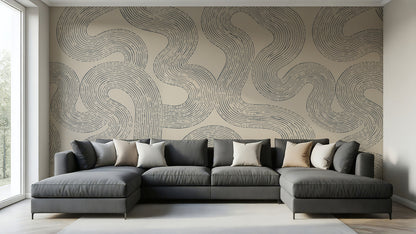 A modern gray sectional sofa with multiple throw pillows is positioned in front of a large wall covered in a beige wallpaper mural with a dark gray wavy line pattern. The room has light wood floors, a white window frame on the left, and a light gray rug on the floor.