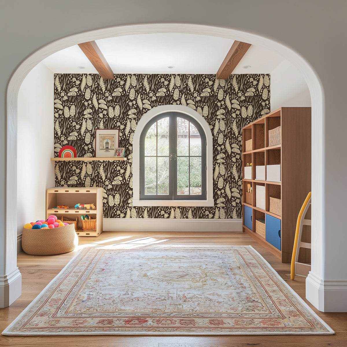 A styled children's playroom viewed through a white arched doorway, featuring dark and beige botanical patterned wallpaper, a large beige area rug, light wood shelving, and a window.