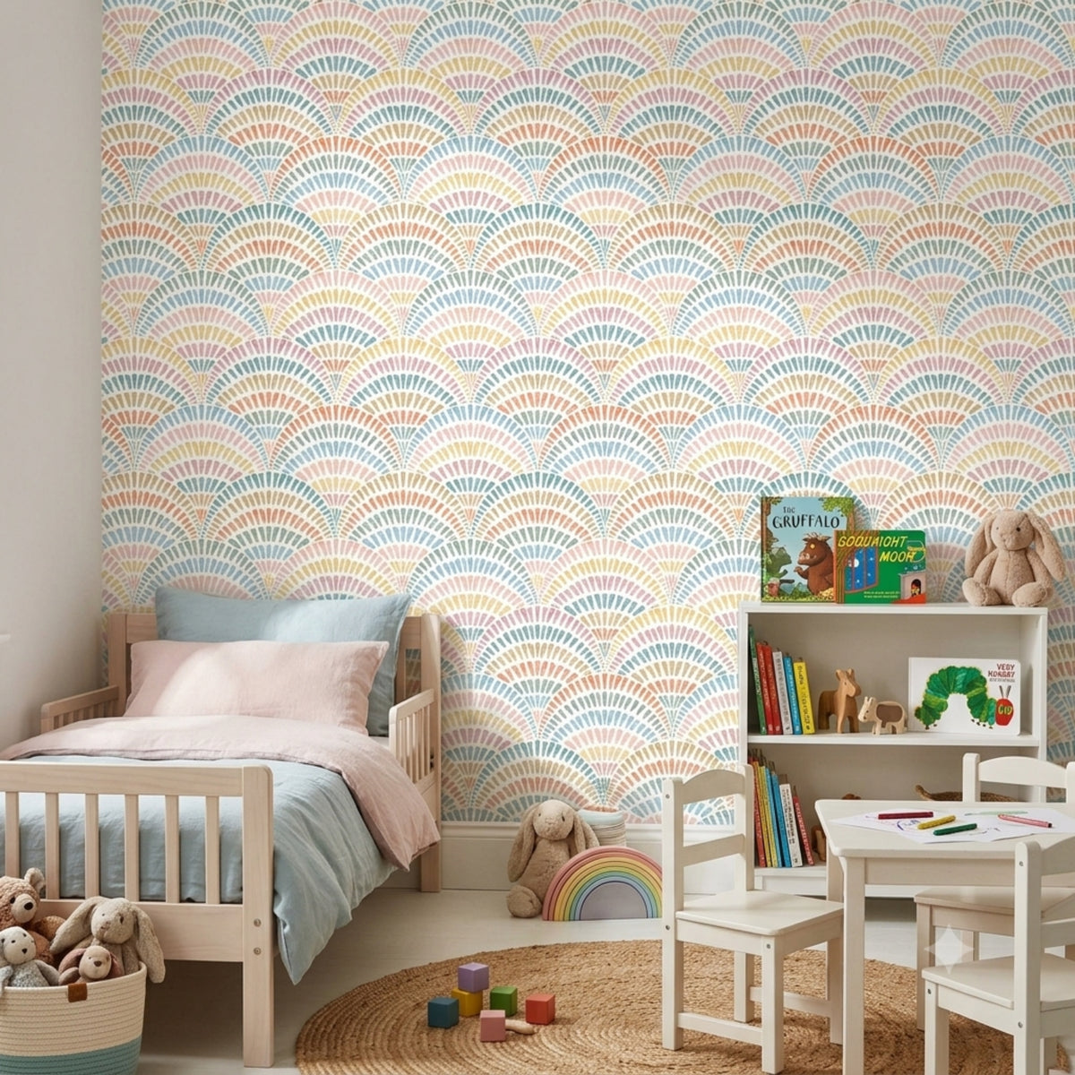 A children's bedroom featuring a wall covered in pastel rainbow scallop-patterned wallpaper. The room includes a light wood toddler bed with pink and blue bedding, a white bookshelf with toys and books, a small white children's table and chairs, a woven rug, and stuffed animals.